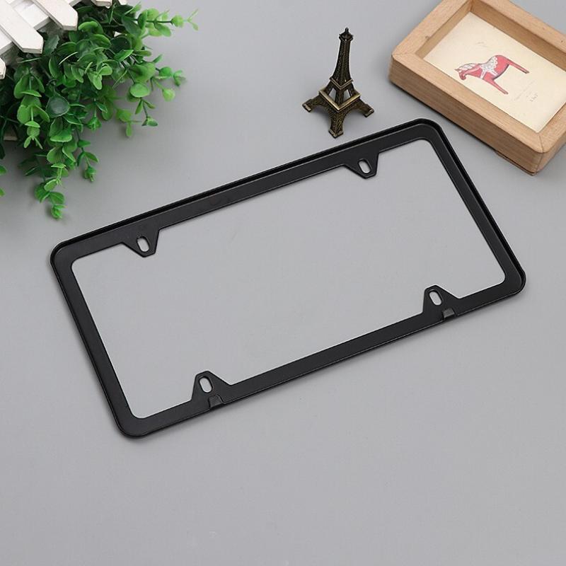 A black stainless steel license plate frame with mounting hardware is displayed on a gray surface next to a small brass objec