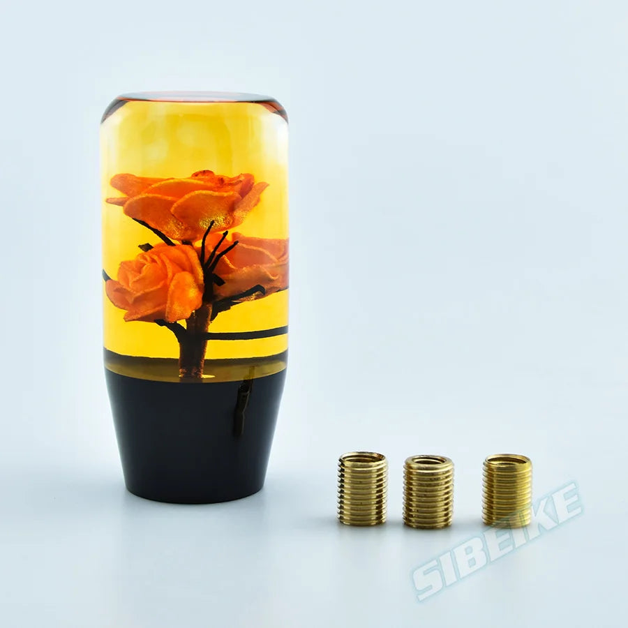 A glass bottle shaped like an orange flower sits on a black base with gold gear shift knobs nearby.