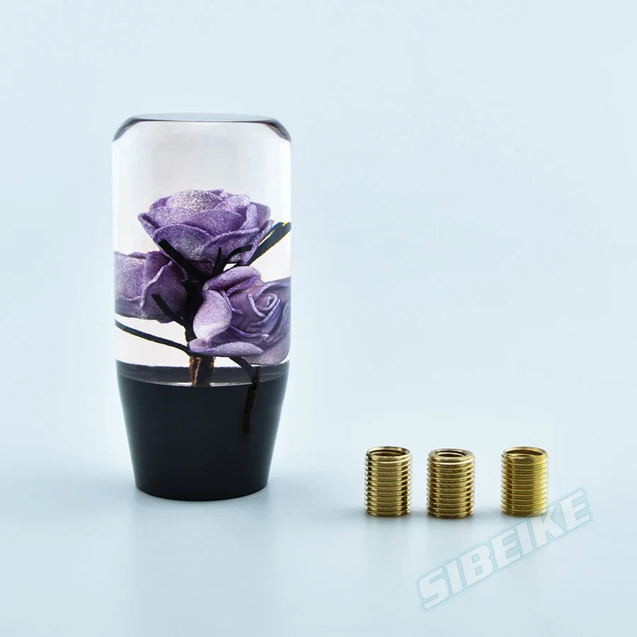 A clear glass cylinder containing a purple rose flower and black base with gold gear shift knob adapters.