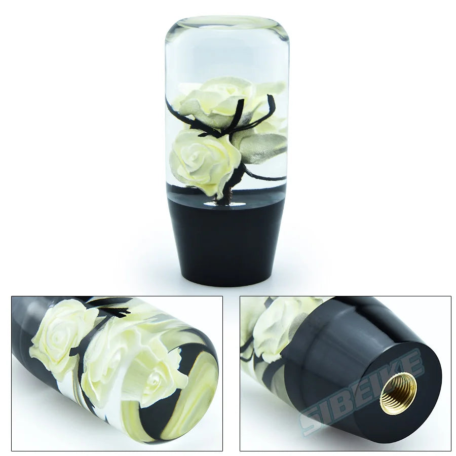 A clear glass container holding a white rose and black leaves, used as a gear shift knob with adapters.