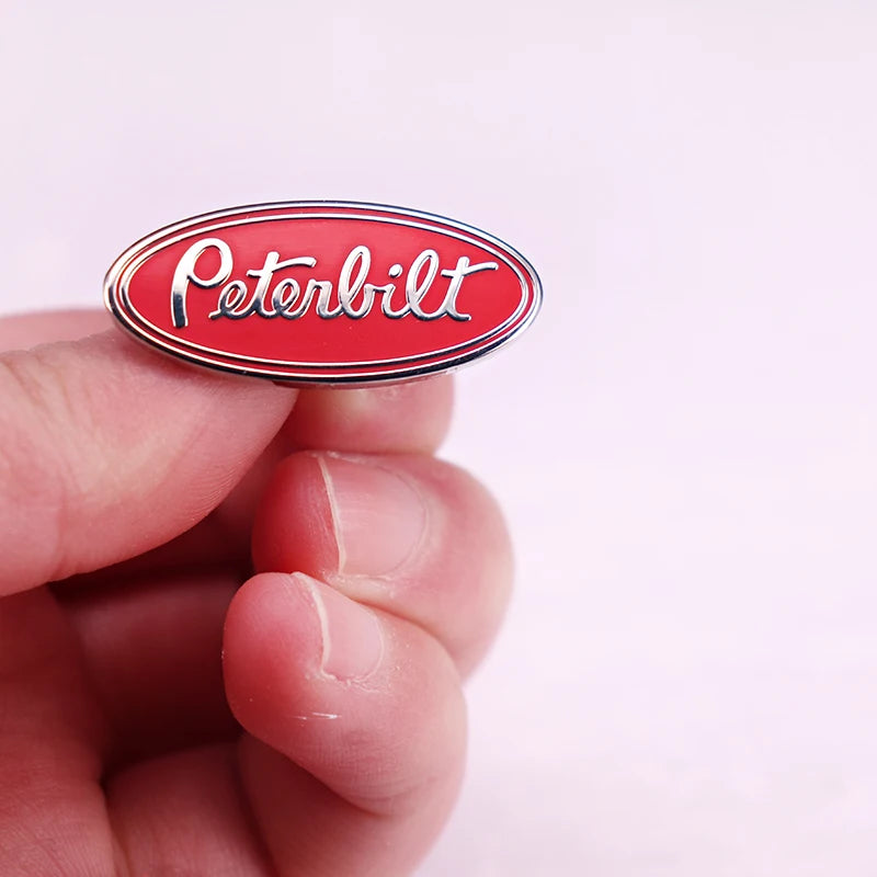 A hand holds a red oval Peterbilt truck emblem pin brooch against a white background.