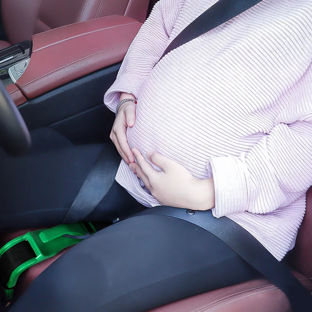A pregnant woman gently holds her belly while seated in a car, wearing a light pink knitted sweater.