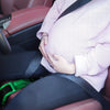 A pregnant woman gently holds her belly while seated in a car, wearing a light pink knitted sweater.