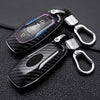 A black carbon fiber car key cover with silver hardware is shown on a dark leather surface.