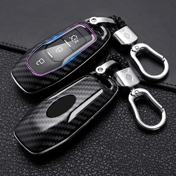 A black carbon fiber car key cover with silver hardware is shown on a dark leather surface.