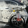 A close-up of a black car being cleaned with white foam from a pressure washer adapter.