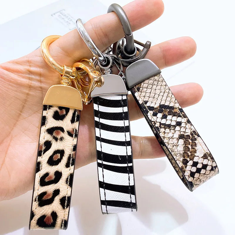 Una mano sostiene un juego de llaveros de cuero con estampado de serpiente leopardo y herrajes de oro y plata.