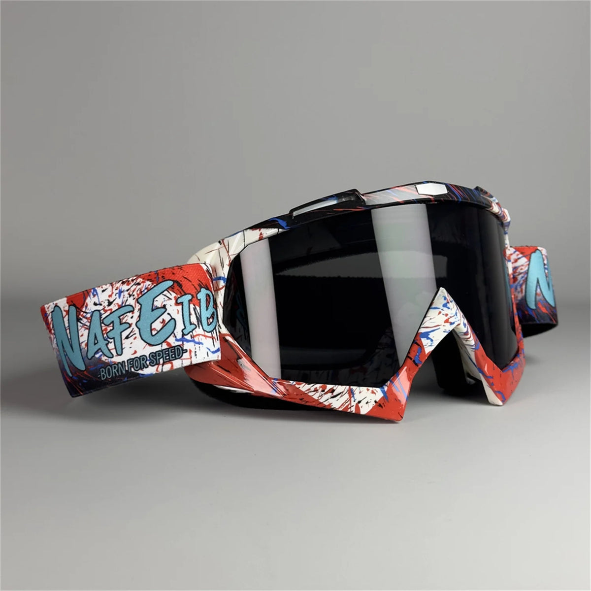 Un par de gafas de motocicleta rojas y blancas con texto azul en el marco, apoyadas sobre una superficie gris.
