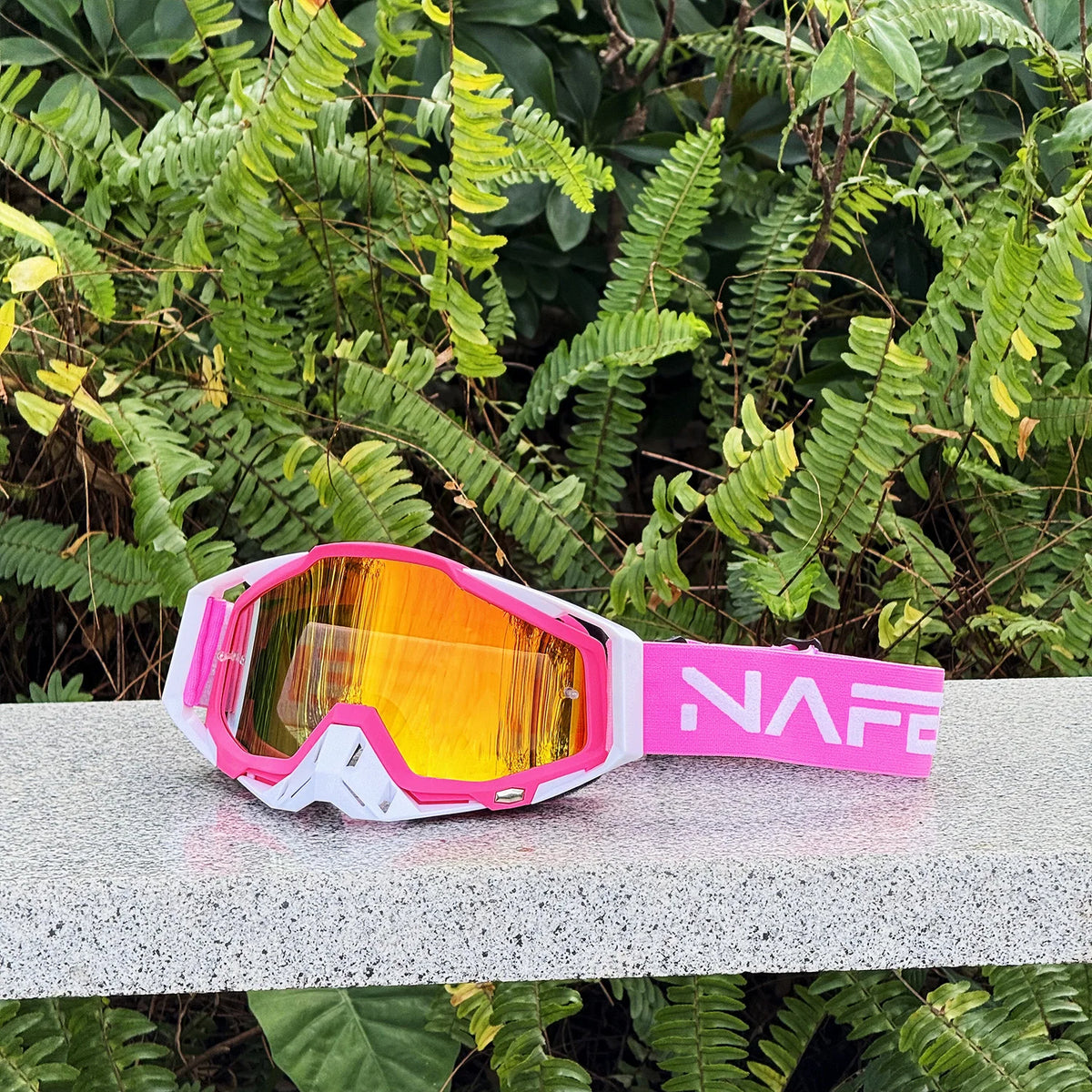Une paire de lunettes de sport pour moto roses et oranges, durable, posée sur une surface claire parmi le feuillage vert.