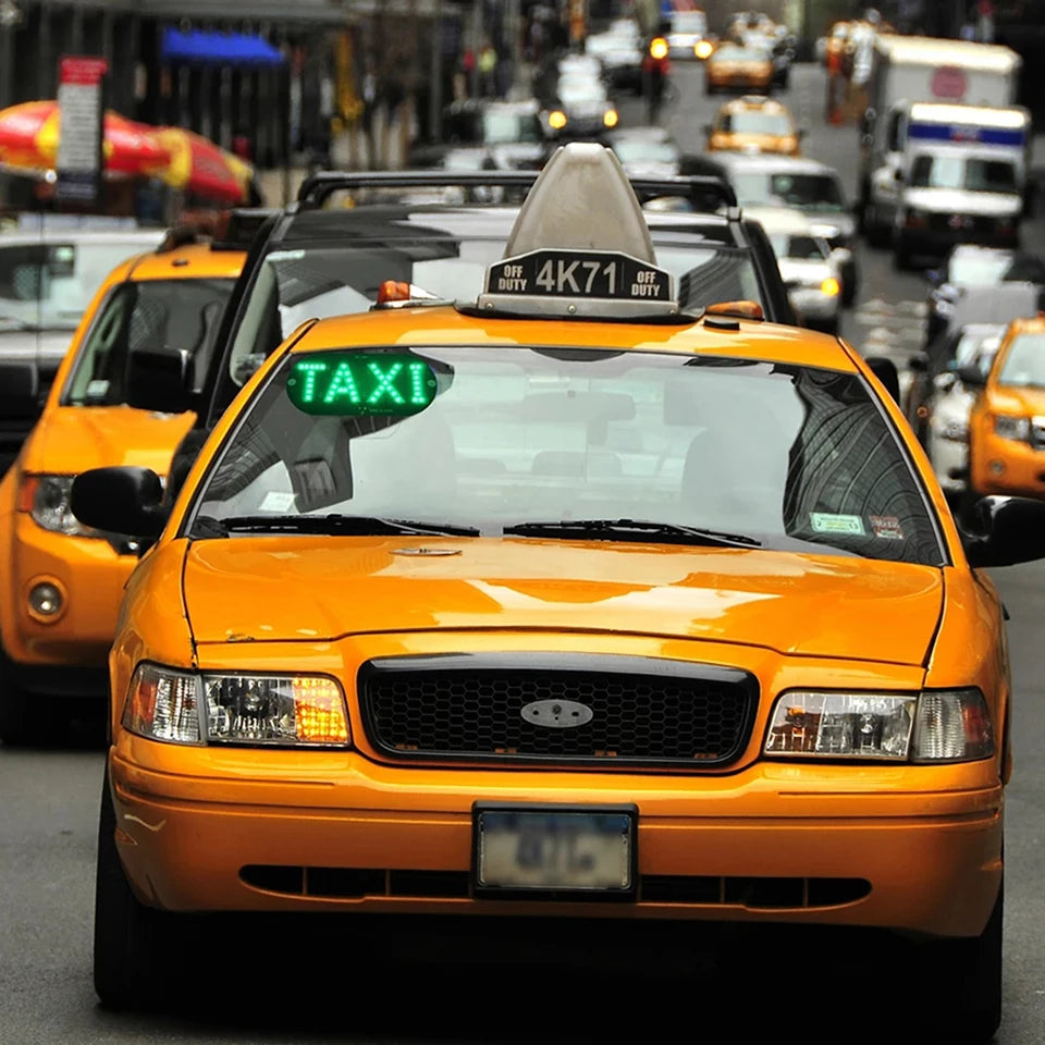 A bright orange taxi cab driving on a city street with an LED indicator light visible on the windshield.