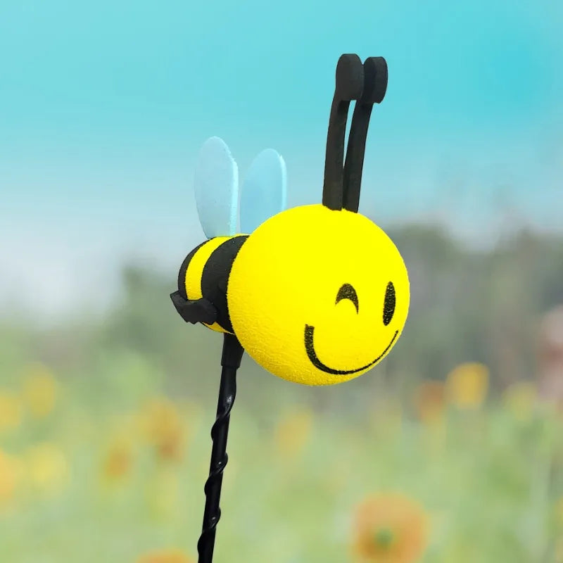 A yellow bee-shaped antenna decoration with blue wings and a black stem against a blurred green background.