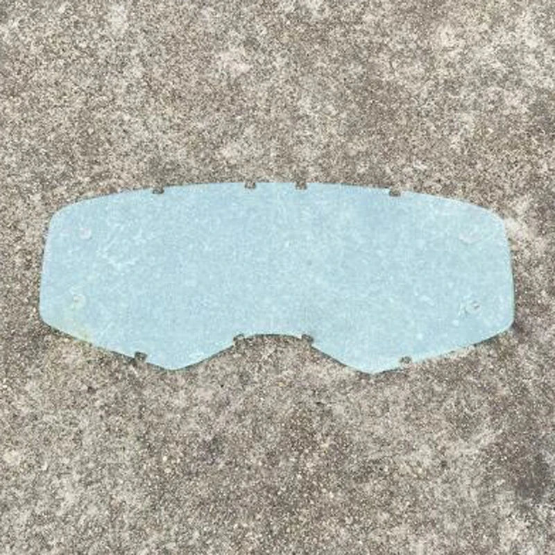 A translucent light blue safety shield or visor for motorcycle or outdoor goggles placed on a gray textured surface.