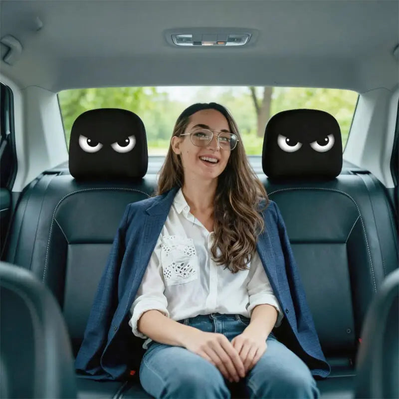 A woman sits in a car, wearing glasses and a blue jacket, with two black angry eye headrest covers visible on the car seats.