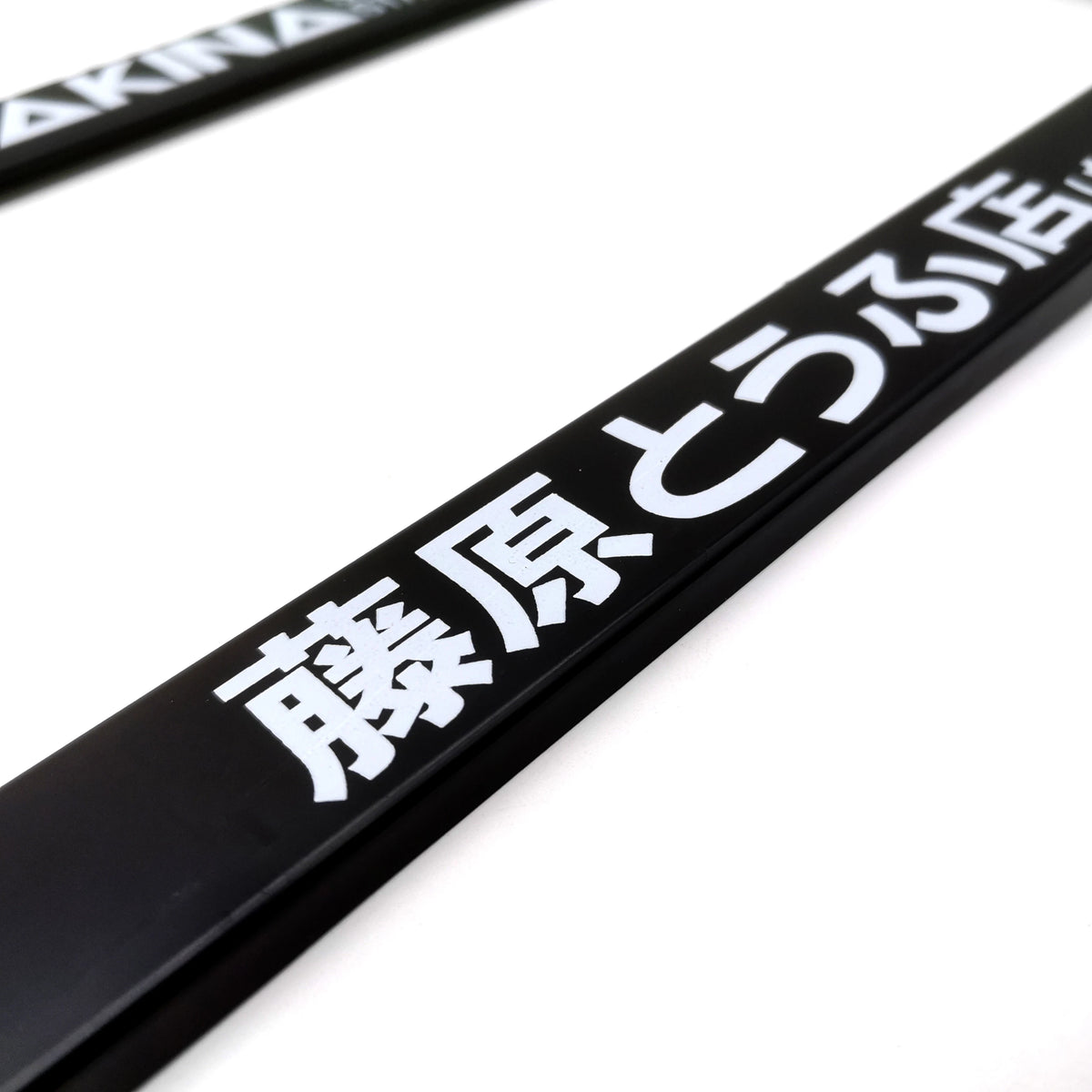 A close-up of a black license plate frame decoration with white Japanese text on a white background.