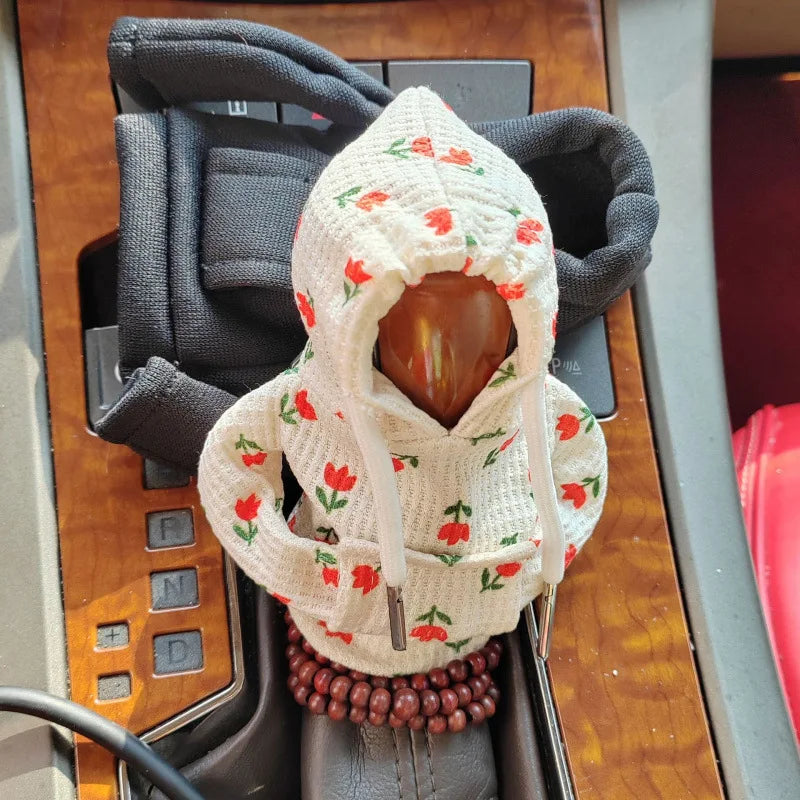 A cozy knit gear shift cover made of cream-colored yarn with red floral accents is shown in a car's gear shift console.