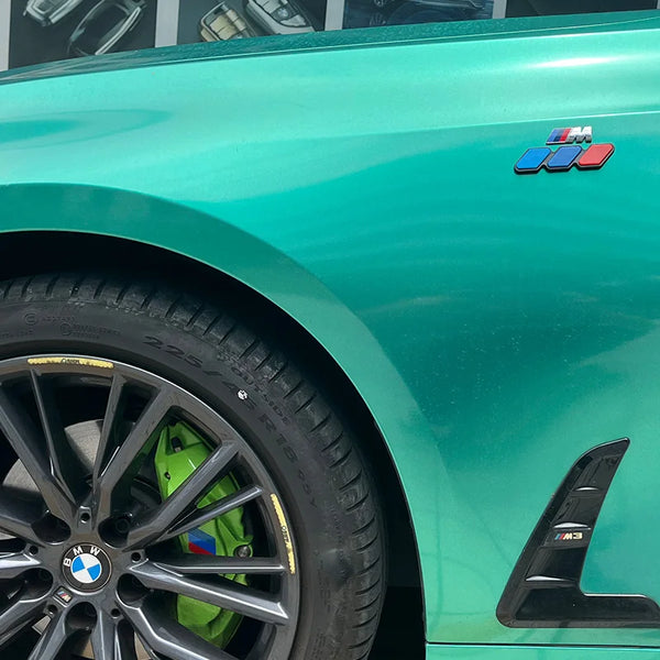 Close-up of a teal BMW fender with a tri-color 3D car grill decal and a black alloy wheel.