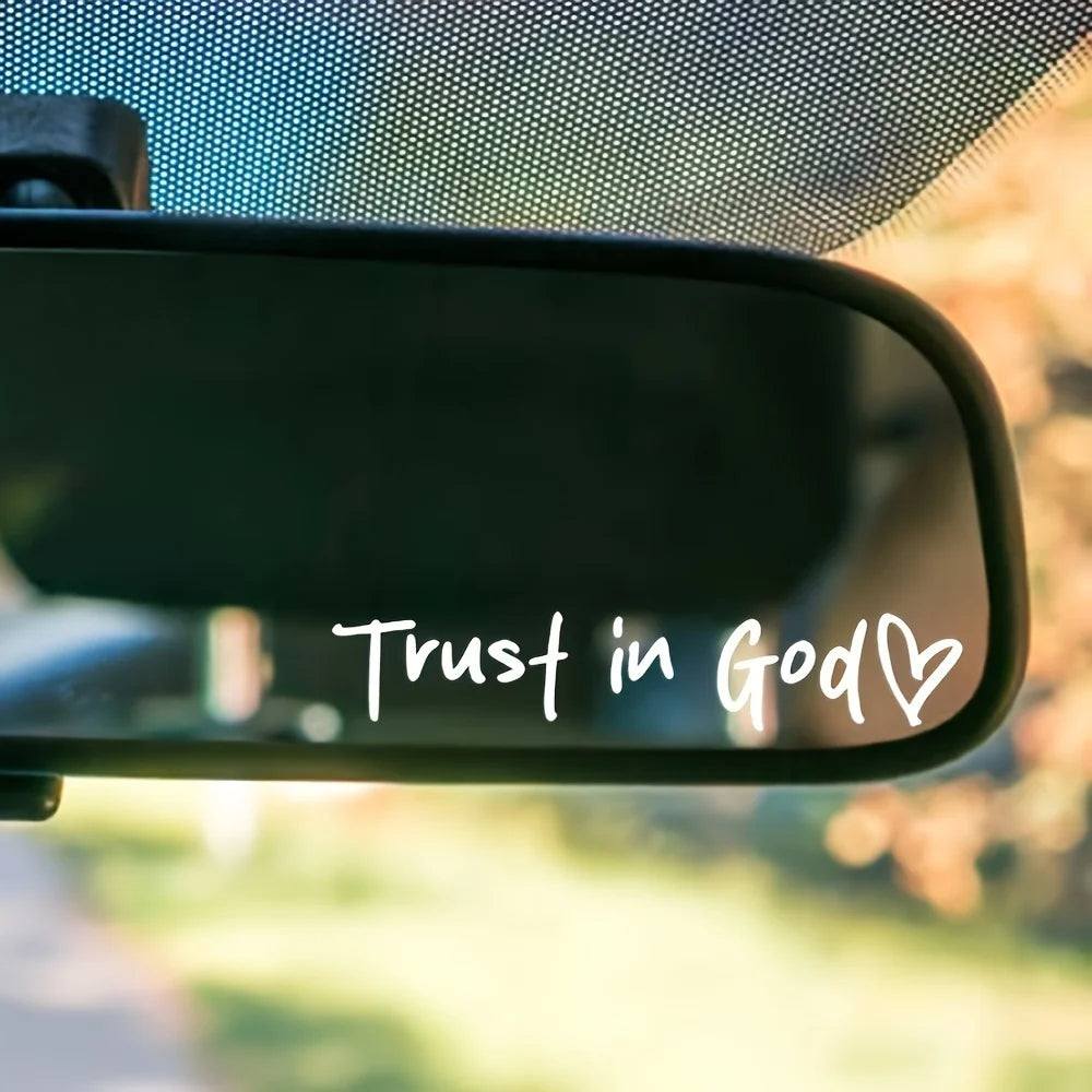 A close-up view of a car rearview mirror with the text Trust in God and a heart decal on it.