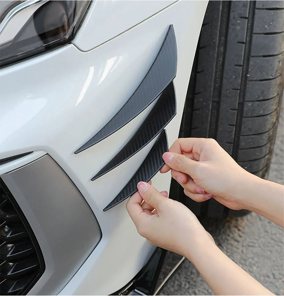 Hands are installing black universal car spoiler fins onto the front bumper of a white car.