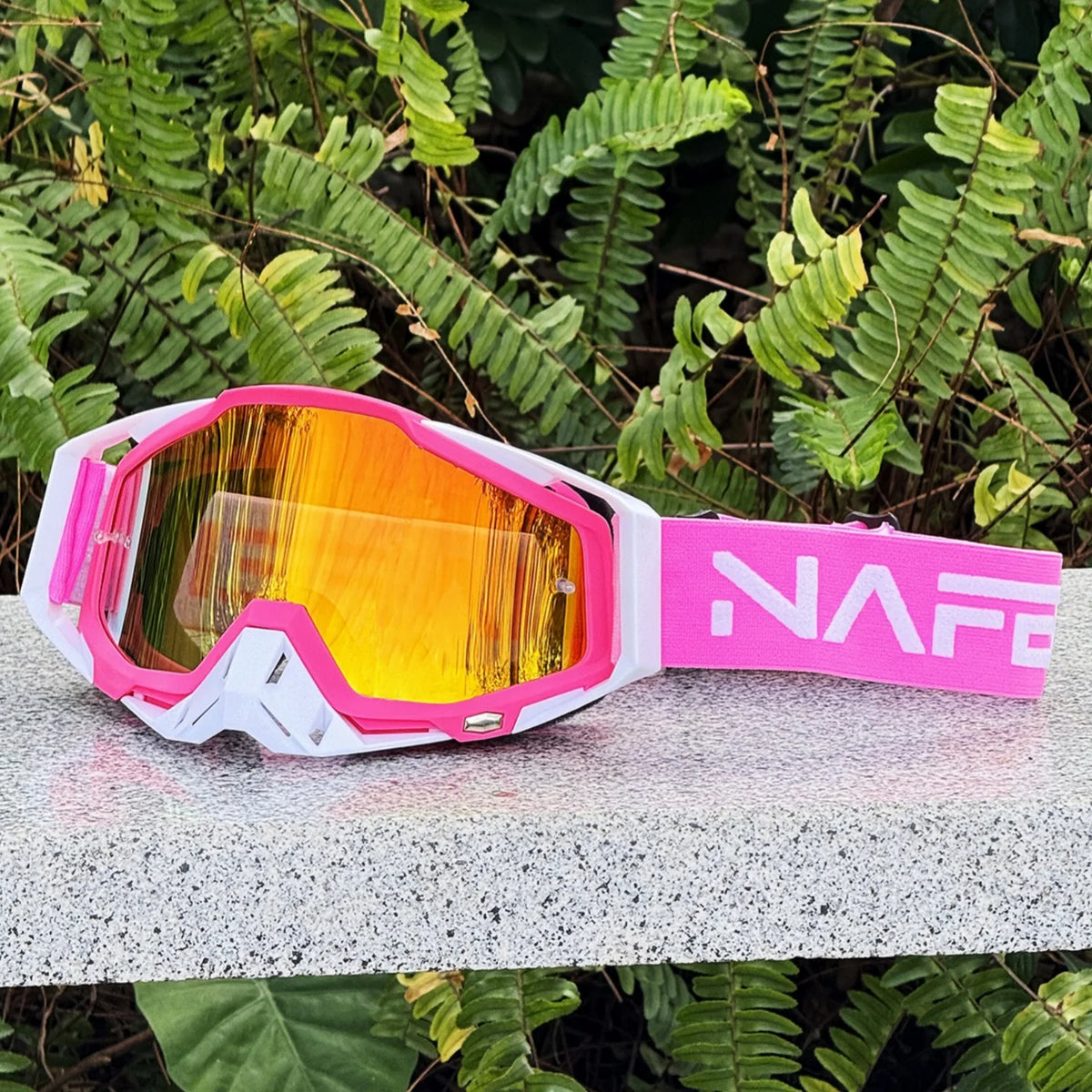 Pinke und weiße strapazierfähige Motocross-Goggles auf grauer Oberfläche mit grünem Laub im Hintergrund.