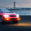 A red Honda Civic sedan with fog lights illuminated drives on a road near a large suspension bridge at dusk.