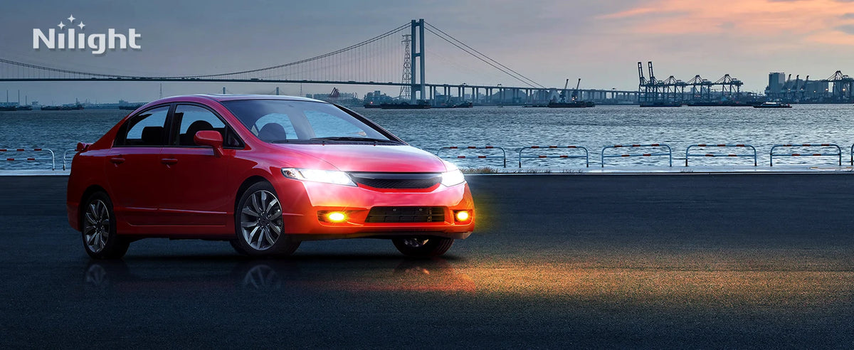 A red Honda Civic sedan with fog lights illuminated drives on a road near a large suspension bridge at dusk.