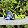 Ein Paar gelb-blaue NAFE Motocross-Goggles auf einer grauen Oberfläche inmitten grüner Vegetation.