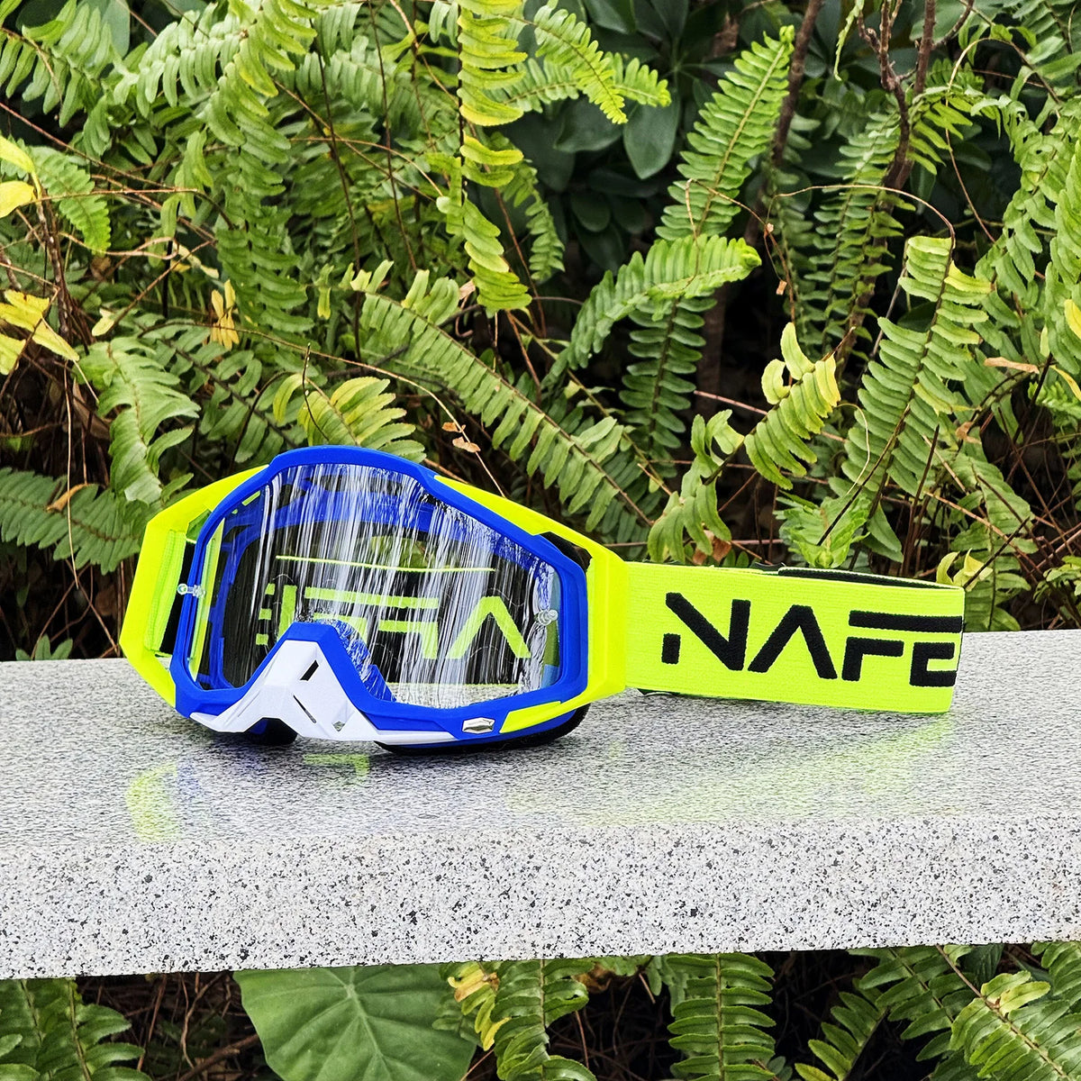 Ein Paar gelb-blaue NAFE Motocross-Goggles auf einer grauen Oberfläche inmitten grüner Vegetation.