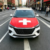 A red and white medical cross flag displayed on the hood of a white car in an urban street setting.