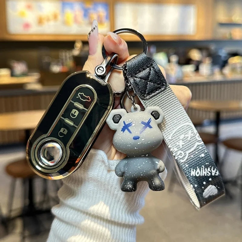Une main tient une housse de clé de voiture en cuir de luxe avec un charm de visage d'ours gris et une clé USB argentée.