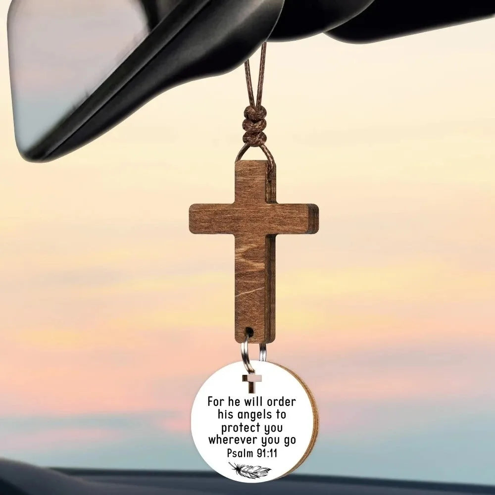 A wooden cross ornament hangs from a black car visor, featuring the text For he will order his angels to protect you wherever