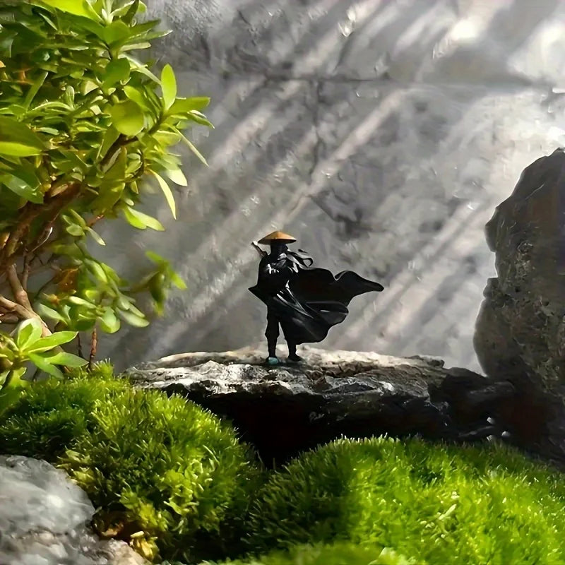A small black samurai statue with a straw hat stands on a mossy rock amidst lush green foliage and sunlight.