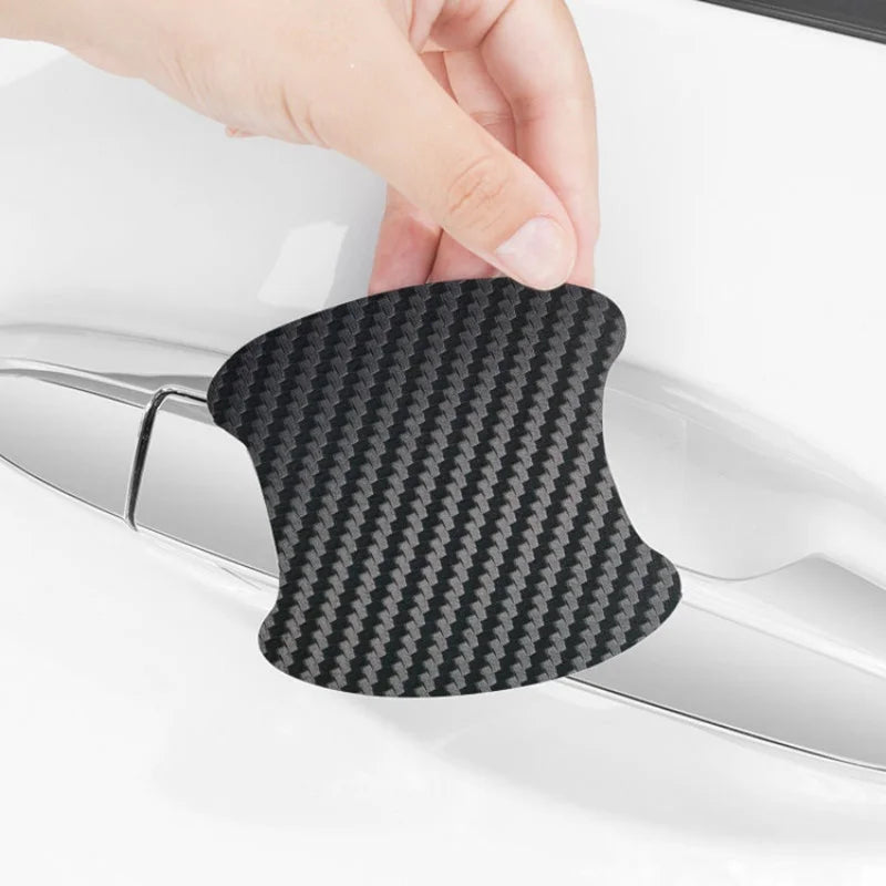 A hand is placing a black carbon fiber door handle protection film onto a car door handle.
