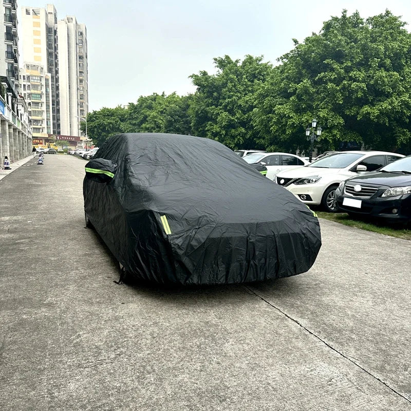 A black universal all-weather car cover is placed on an asphalt road next to parked cars and green trees.