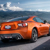 An orange Ford Mustang sports car driving on a scenic road with mountains in the background.