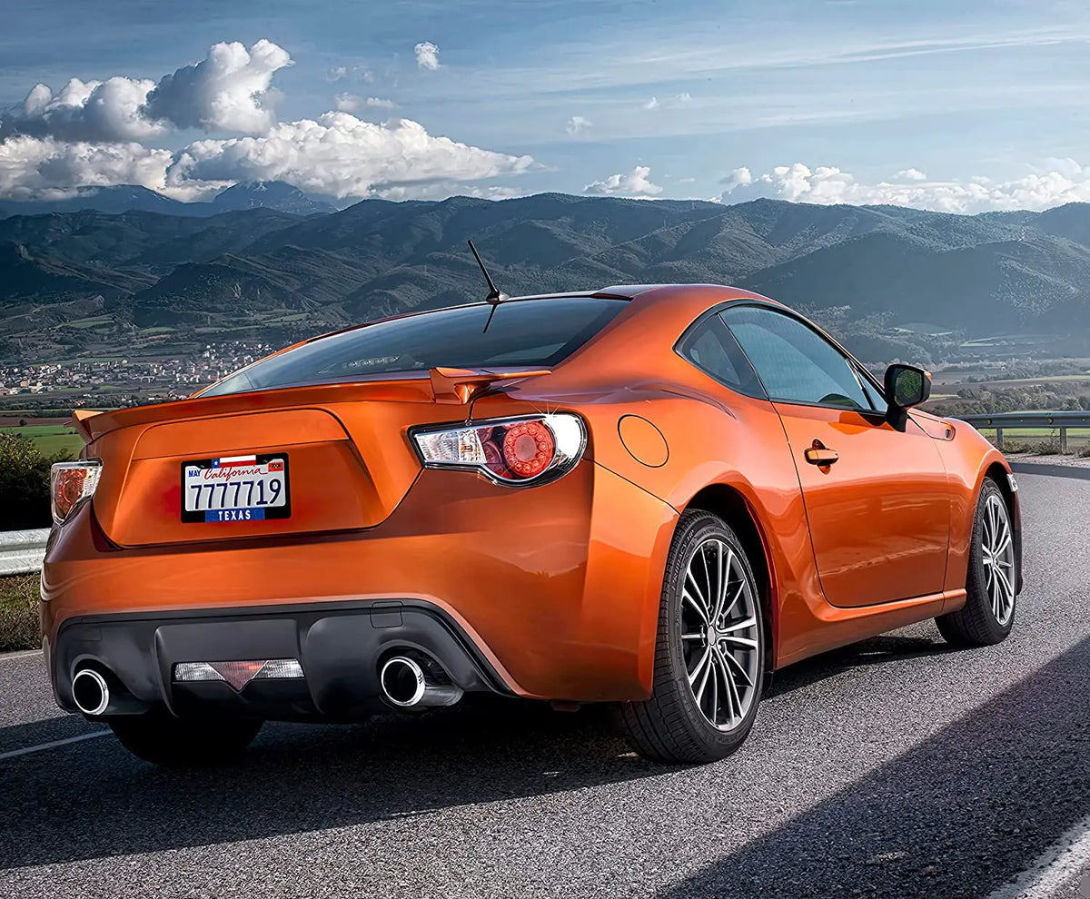 An orange Ford Mustang sports car driving on a scenic road with mountains in the background.