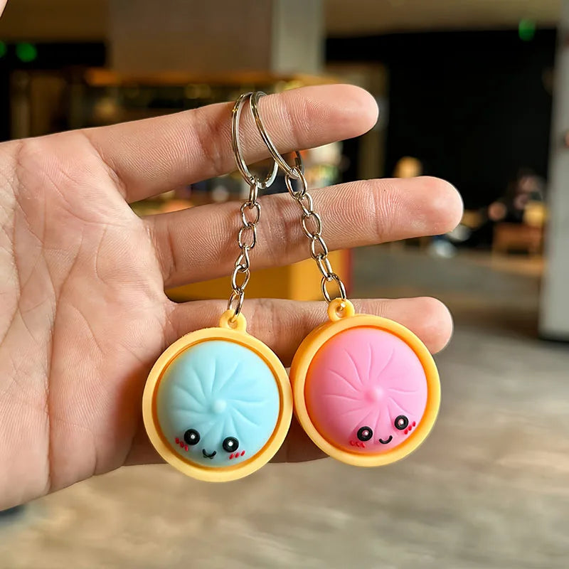 A hand holds two yellow silicone keychains with pink and blue faces and silver jump rings.