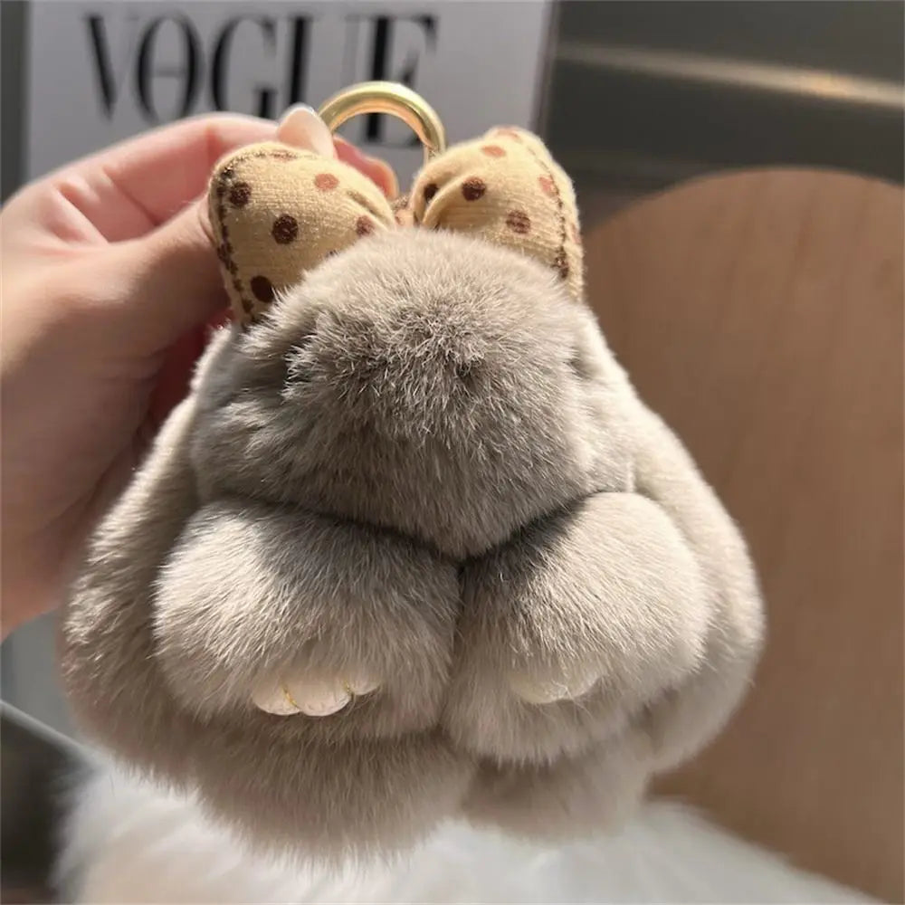 A close-up of a plush bunny keychain with soft gray fur and gold ring being held by a hand.