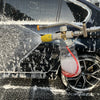 A hand is using a dual-outlet pressure washer foam cannon adapter to spray soapy water onto the exterior of a dark car.