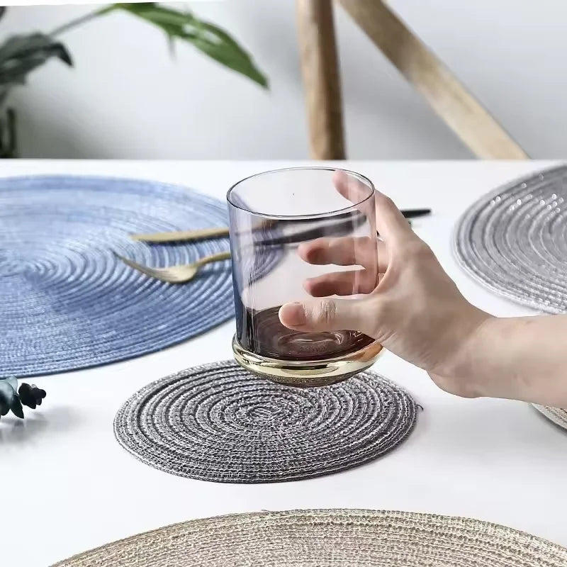 A hand pours dark liquid from a glass onto a round gray placemat on a white table.