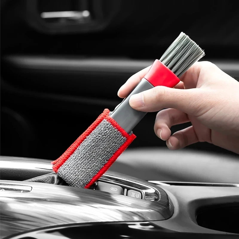 A hand is using a red and gray brush to clean a car air vent.