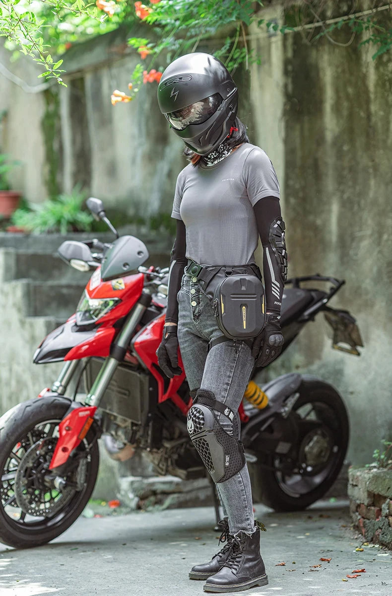 A person wearing a black motorcycle helmet and black gloves stands next to a red motorcycle with a black fanny pack.