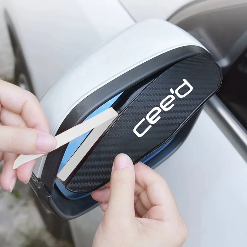 A hand is removing a carbon fiber rain shield from a white car's rearview mirror housing.
