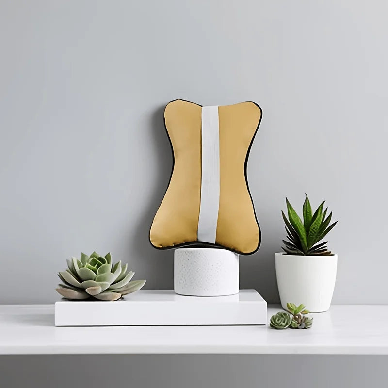 A beige ergonomic auto headrest cushion with a white strap is displayed on a white stand next to a potted succulent plant.