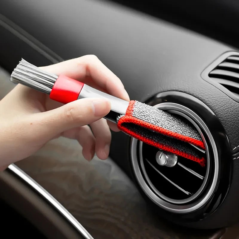 A hand is using a red and silver brush to clean a car air vent grille.