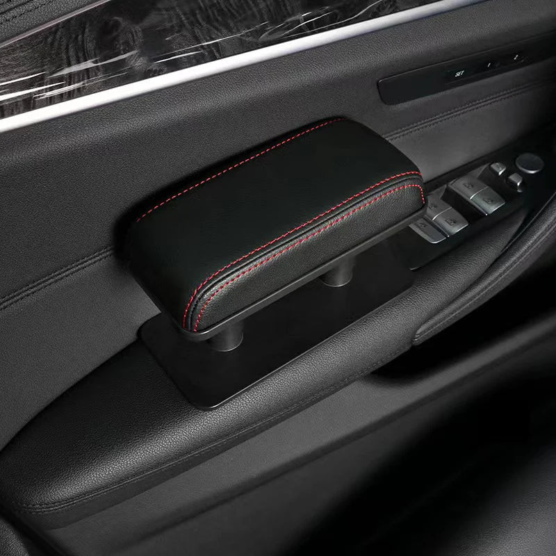 A black ergonomic vehicle armrest cushion with red stitching is shown in a car interior.
