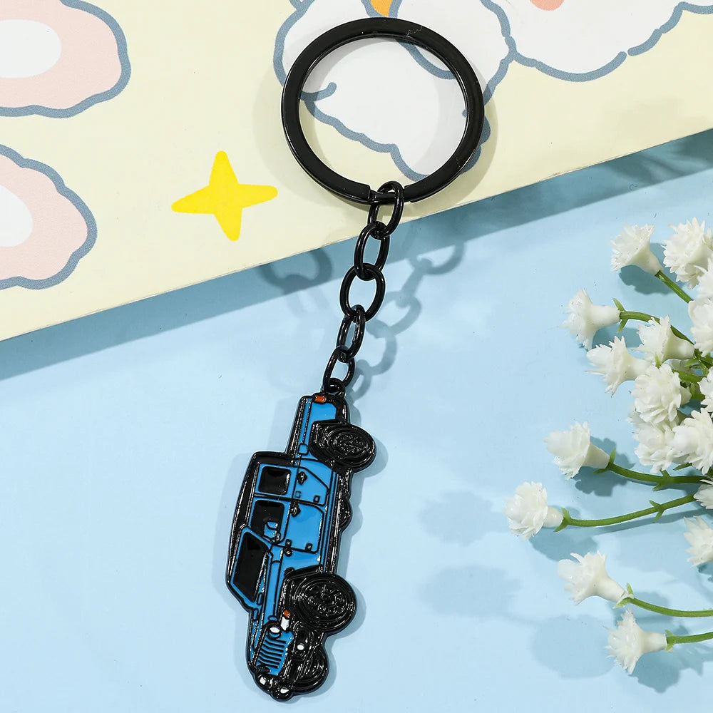 A black keychain shaped like a blue classic alloy car, resting on a light yellow background with pastel clouds.