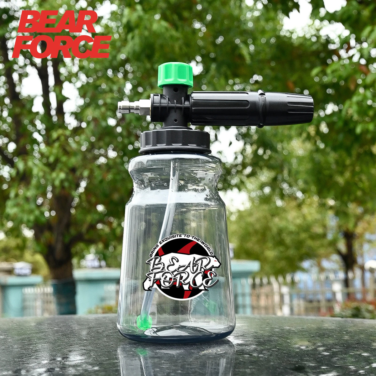 A clear glass bottle containing a foam lance with a green nozzle and black handle, set against an outdoor background.