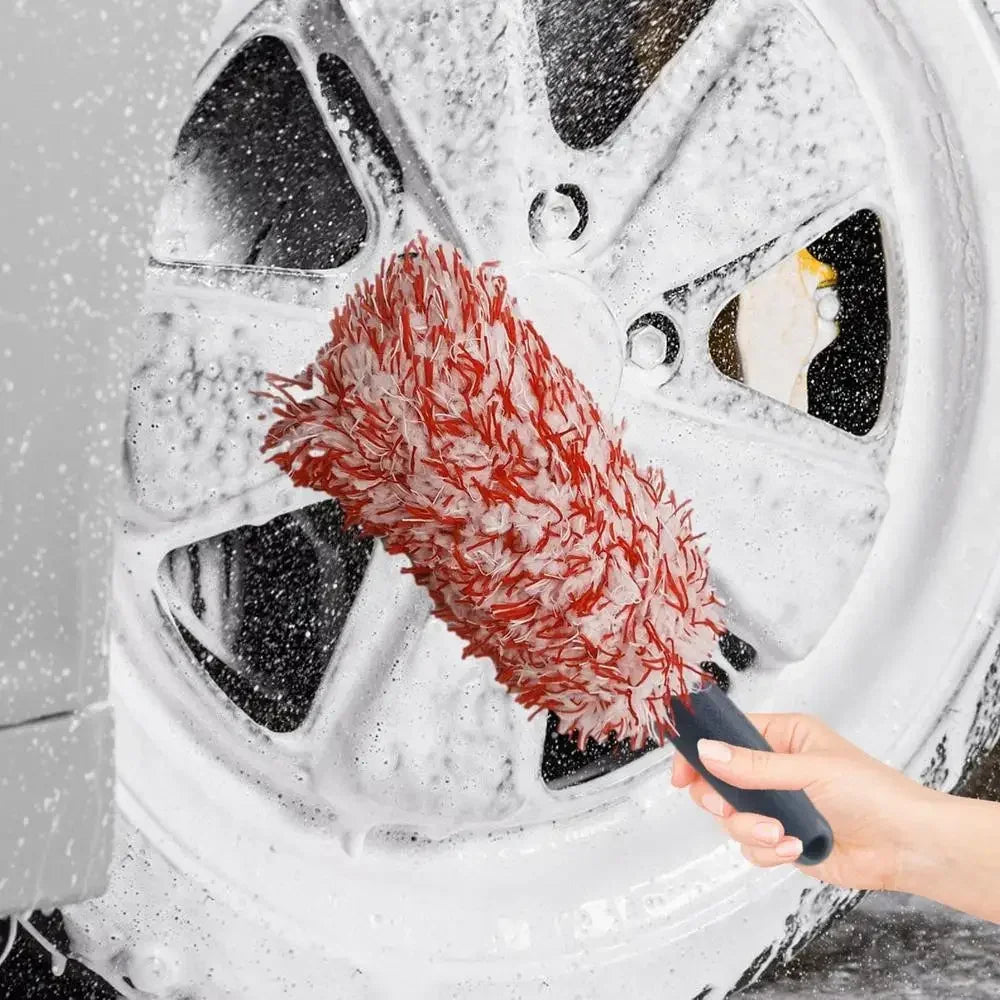 Une main utilise une brosse en microfibre rouge pour nettoyer un jante de pneu blanc avec de l'eau savonneuse.