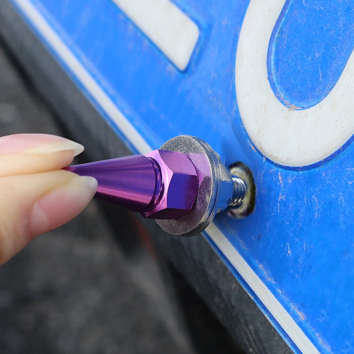 A hand holds a purple M6 license plate fixing screw against a blue automotive surface.