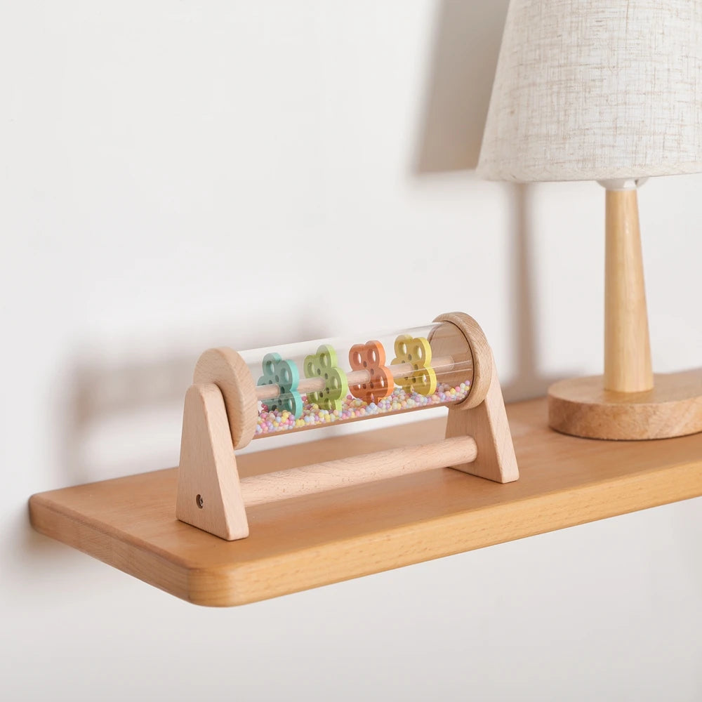 A wooden rainmaker musical toy with colorful plastic raindrops is displayed on a light wood shelf next to a lamp.